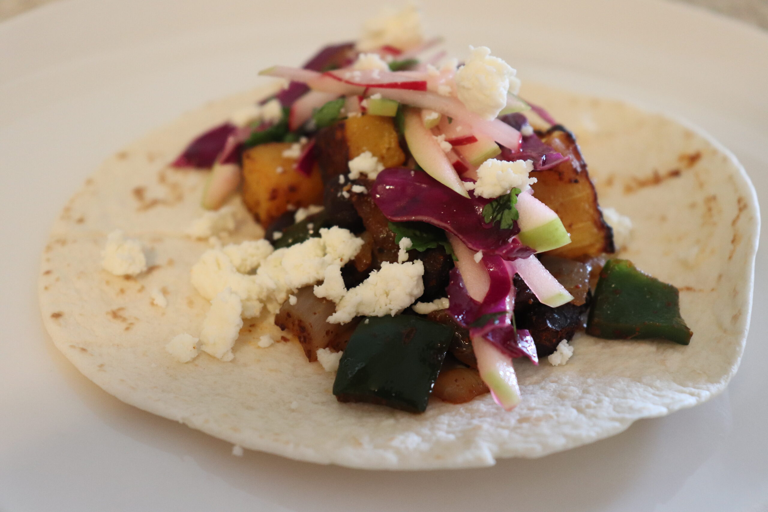 Taco shell topped with a vegan filling of butternut squash, poblano peppers, onions, and black beans topped with a slaw made of purple cabbage, raddish, and apples.